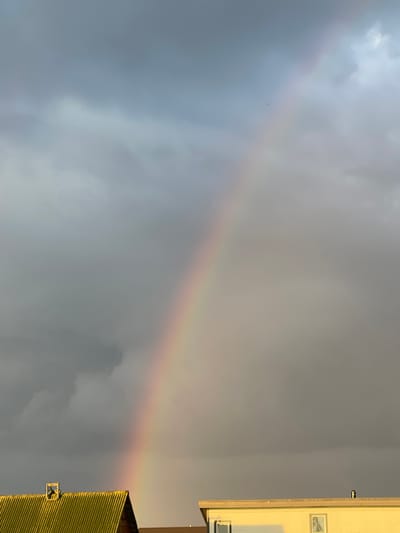 The image shows a partially visible rainbow against a cloudy sky. In the foreground, the roofs of two buildings are visible. The building on the left has a yellow, corrugated metal roof with a small wooden structure on its peak. The building on the right is lighter in color and has windows. The scene is likely an outdoor view from a residential area in Zaķumuiža, Latvia, possibly after a rain shower given the presence of the rainbow.
