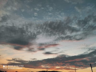 This media file depicts a sunset scene over Mliiv, Ukraine. The sky is filled with dramatic clouds, ranging from dark, ominous grey to lighter, wispy formations tinged with pink and orange hues from the setting sun. The overall impression is one of a dynamic and perhaps turbulent sky.
The lower portion of the image shows a dark silhouette of a landscape, likely hills or a treeline, against the vibrant sky. There are also two vertical poles visible in the foreground, suggesting the presence of human infrastructure such as power lines or some form of antennae. The light indicates it is either sunrise or sunset, with the warm colors predominantly in the lower sky fading into cooler blues and greys higher up.
Visible text in the image reads "REDMI NOTE 9 PRO" and "AI QUAD CAMERA", indicating this is a photograph taken with a mobile device and includes a watermark. The scene evokes a sense of natural beauty and the power of the elements, with a hint of human presence. The emotional tone is somewhat dramatic due to the cloud formations and the contrast between the dark silhouettes and the colorful sky.