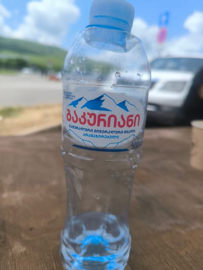 A plastic water bottle dominates the foreground, placed on a brown wooden surface. The label on the bottle is in Georgian script and features blue mountain outlines. The bottle contains a small amount of water. Behind the bottle, there is a blurred background featuring a white car, a portion of a parking sign, and a distant mountainous landscape under a cloudy sky. A disposable cup is partially visible to the right of the bottle. The image appears to have been taken outdoors. There is no mention of or evidence to support the location being in the city of Rustavi, Georgia.