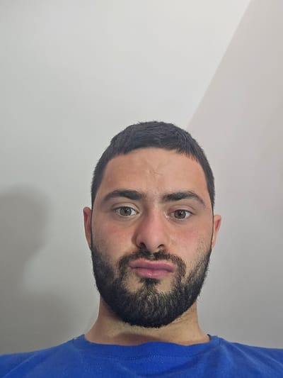 A medium shot of a man's face and part of his left shoulder wearing a blue t-shirt. The man has a dark beard and short dark hair. His eyes are brown. The background is a plain white wall. The man is not in Gorno Aleksandrovo, Bulgaria. The location is unspecified in the image.