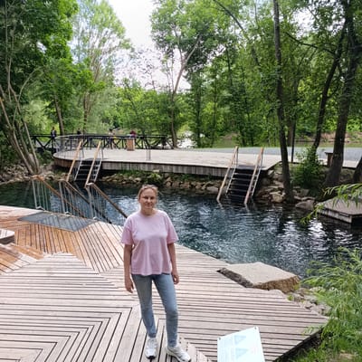 A woman stands on a wooden boardwalk by a clear blue body of water surrounded by lush green trees. She is wearing a light pink t-shirt, blue jeans, and white sneakers. The boardwalk features a geometric pattern of wooden planks. In the background, there are stairs leading down into the water, and a larger wooden walkway with railings, where several other people are visible. Some people appear to be walking on the walkway, while others are near the water's edge. A distant bridge can be seen in the background. The water is calm with ripples reflecting the light. The scene appears to be a natural spring or swimming area. The time of day seems to be daytime, with ample natural light, suggesting fair weather. The overall atmosphere is peaceful and recreational. A sign with some text and symbols is visible in the lower right corner of the image. The location is Sdt Shcherbakovka, Russia.