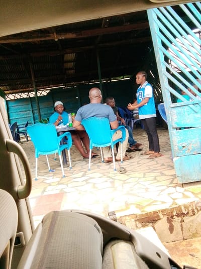 The image depicts an outdoor or semi-outdoor social gathering within an establishment in Ore, Nigeria. The scene is observed from the interior of a vehicle, with a tan-colored seat and armrest visible in the foreground.

Within the establishment, a corrugated metal roof supported by wooden beams covers a paved area. The walls are constructed from horizontal blue wooden planks. Five individuals are present. Three are seated around a dark-colored table on bright blue plastic chairs with white legs. One woman, wearing a light blue top and a white cap, sits to the left of the table, looking left. A bald man, with his back to the camera, wears a dark grey t-shirt and light-colored trousers, seated on a blue chair. Another man is partially visible to his right. On the table, a green bottle and other small items are discernible.

A person, appearing female, stands to the right, facing the seated group. She wears a light blue and white top with a number "18" (or letter "B") and black trousers, with her hands clasped in front. Her footwear consists of flip-flops. To the far right, a blue wooden louvered gate or partition is present. The floor is paved with light-colored, irregularly shaped mosaic tiles. Overhead, a single light fixture hangs from the roof structure. The overall environment suggests an informal setting for dining or conversation.