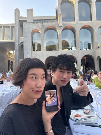 In Esplugues de Llobregat, Spain, at a formal outdoor dining event set against a strikingly modern architectural backdrop with arched windows and concrete structures, two individuals are the primary focus. A woman with short, dark hair and bangs is positioned on the left, holding a smartphone displaying a picture of a small dog with brown and white markings. She is looking to her left with a slight smile. To her right, a person with dark, curly hair is pointing towards the phone with their right hand, looking towards the viewer. Both are dressed in dark attire. The table is set with white tablecloths, fine china, cutlery, and glassware. In the background, other guests are seated at tables, and more of the unique architecture is visible.