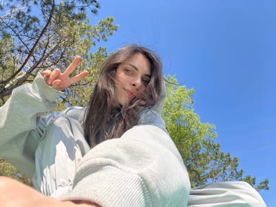 This is an outdoor selfie featuring a young person with long, dark brown hair, looking directly at the camera. They are wearing a light gray or mint green hooded sweatshirt with the hood up. Their light-colored eyes are clearly visible, and their expression is neutral to slightly smiling. The person's right hand is raised, making a peace sign gesture with two fingers extended, and a ring is visible on one finger.
The photograph is taken from a low angle, with the camera pointed upwards towards the subject and the sky. The background showcases a bright, clear blue sky, indicating a sunny day. Green tree foliage, including pine trees with their distinct needles and cones, as well as deciduous trees with fresh, lighter green leaves, frame the edges of the sky. Faint, thin lines, possibly power lines, can be seen against the blue sky in the upper left.
The setting appears to be a natural, outdoor environment, likely a park or a wooded area, consistent with the specified location of Слизнево, Russia. The overall mood is casual and relaxed, captured during bright daylight. There is no visible text in the image.