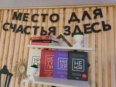 The image shows a close-up of a wall decorated with wooden slats. A black banner with black letters strung across the slats reads "МЕСТО ДЛЯ СЧАСТЬЯ ЗДЕСЬ", which translates to "A PLACE FOR HAPPINESS HERE". Below the banner, a white shelf displays four books. The books are of different colors: white, red, purple, and black, each with a title in white text. The white book has the Cyrillic letters "НИ" and "СЫ" on it. The red book has "НЕ" and "ТУ" and "ПИ". The purple book has "НИ" and "ЗЯ". The black book has "НЕ" and "НОЙ". To the left of the books, a small decorative white figure with a green top sits on the shelf. Further to the left, a small golden sunburst mirror is mounted on the wall. To the far left, a black coat rack or hanger is visible. The scene appears to be indoors, possibly in a home or small business, in Slavyansk-na-Kubani, Russia.