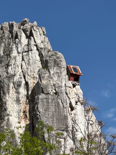 A weathered, rusted metal box with a solar panel attached sits precariously on a rocky cliff face. The box is red on one side and has an open section that reveals a dark interior. The cliff is layered and composed of light gray stone with textured surfaces. Some small plants grow from the rock. Bare branches and a tree with green leaves appear at the top-right edge of the frame. The background is a cloudless blue sky. The image was taken at Milanovo, Bulgaria.