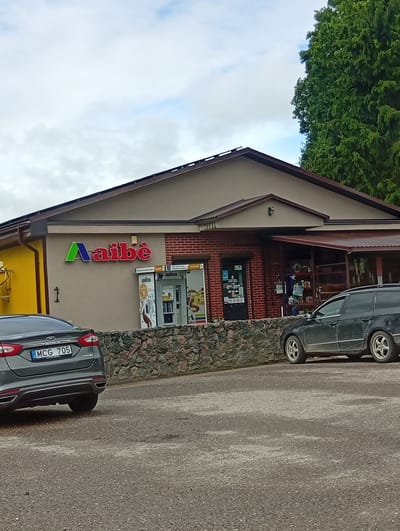 The image shows the exterior of a "Aibė" convenience store in Šaukėnai, Lithuania. The store has a light brown facade with a darker brown trim and a pitched roof. A prominent "Aibė" logo, with green, blue, and red lettering, is displayed on the facade. To the right of the logo, there are two Lavazza coffee machines. The store is built with some brickwork around the entrance and windows. A low stone wall runs along the front of the building. Two cars are parked in the foreground on a gravel lot. The car on the left is a dark gray Ford Fusion with a license plate reading "MCG 705". The car on the right is a dark colored station wagon. A large green tree is visible on the right side of the image, and the sky is overcast with some blue patches. The scene appears to be a typical daytime view of a local shop.