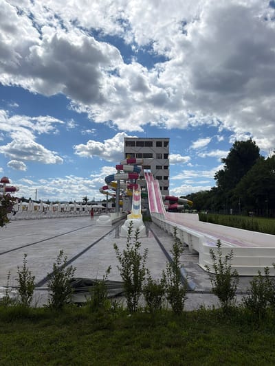 In Ivanyane, Bulgaria, an outdoor water park features a towering multi-story white building with visible windows and a grid-like facade. Attached to the building is a complex of colorful water slides, including spiral sections in red, yellow, and blue, and a long, wide pink and white slide that extends horizontally across the foreground. The ground level is a vast, light-colored concrete expanse with black grates and lines, dotted with small green bushes and patches of grass. In the background, rows of white lounge chairs and umbrellas are neatly arranged under a bright, cloudy sky with patches of blue. Trees form a dense line along the right side of the frame, and a few individuals are visible in the distance, one walking in the pool area and others standing near the tree line. The scene captures a sunny day at the water park.