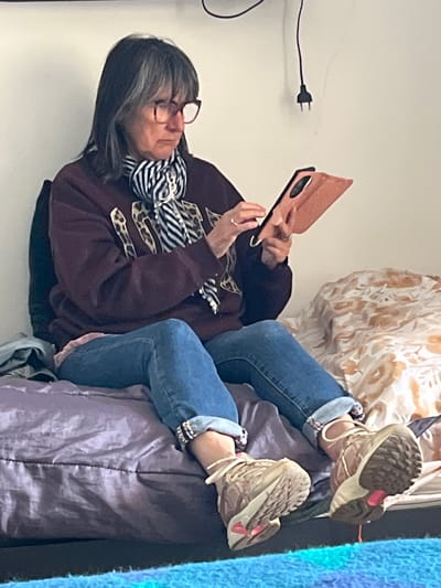 A woman is reclining on a cushion or a bed, looking at a tablet. She is wearing glasses, a maroon-colored sweatshirt with leopard print designs, a striped scarf, blue jeans with the cuffs rolled up, and sneakers. The setting appears to be indoors, with a white wall behind her, a tan and white floral pattern above her, and a blue textile to her right. A power plug is visible on the wall behind the woman. The location in Palavas-les-Flots, France is not evident in the image.