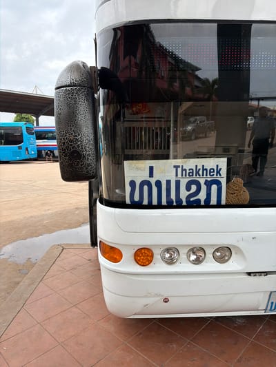 A close-up, low-angle shot shows the rear of a white tour bus parked outdoors at a bus station in Phakhao, Laos. The bus has orange turn signals and multiple clear circular lights on its rear bumper. A sign with blue text, reading "Thakhek" in English and Lao script, is affixed to the back window. To the right of the sign is a brown, textured object, possibly a decorative item or a piece of luggage. The bus's rear view mirror, with a black, cracked, textured surface, is visible on the left side of the frame. In the background, a blue and a red-and-white bus are parked under a covered area. The ground is paved with reddish-brown square tiles, with a wet patch visible on the concrete edge to the left. The sky is overcast.