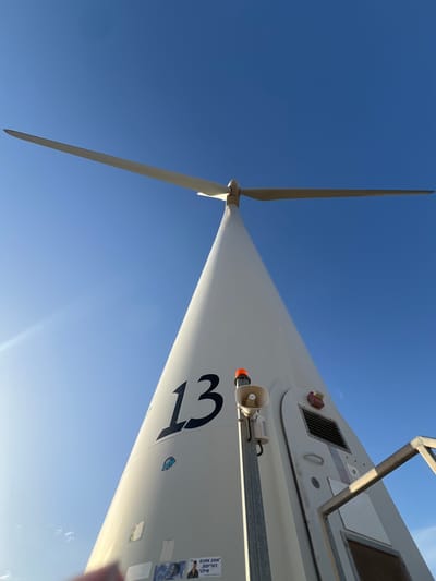 A low-angle shot of a wind turbine against a clear blue sky. The turbine, marked with the number "13", appears to be located in Emek HaMaayanot Regional Council, Israel. The blades are visible, and a speaker is mounted on the turbine structure. Some stickers and labels are on the side of the turbine.