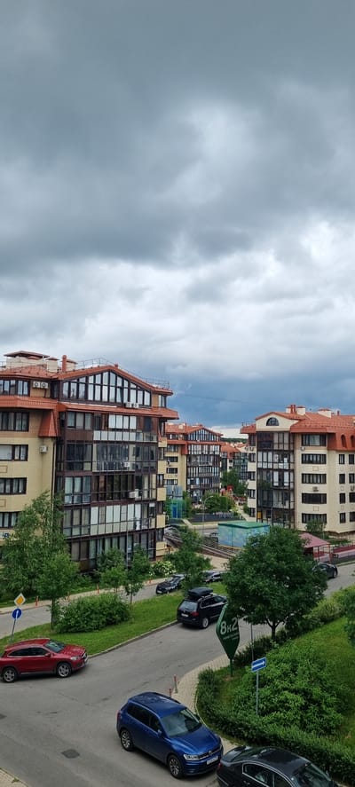 The image depicts a residential street in Romashkovo, Russia, on a cloudy day. The primary subjects are modern apartment buildings with red tiled roofs and large, multi-pane windows. Several cars are parked along the asphalt road, including a red hatchback, a blue hatchback, and a few dark-colored sedans. Lush green trees and bushes line the street and are interspersed between the buildings, suggesting a well-maintained urban or suburban environment.

The scene appears to be a typical residential setting, with no people visible. The overall mood is calm and ordinary, with the cloudy sky hinting at potential rain or overcast conditions, though no rain is actively falling. The architecture of the buildings suggests a relatively new development. A green sign with white text is visible, displaying "8K2" and some indecipherable Russian text. A blue directional sign with a white arrow pointing to the right is also present. There is no specific activity or event taking place; it is a snapshot of a quiet street. The time of day is not explicitly discernible but appears to be daytime.