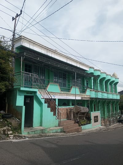 A two-story, light green building stands on the roadside. The upper level features a row of arched columns, windows with bars, and a metal-roofed balcony with a staircase. The lower level is accessed by stone steps, and a closed door is visible on the left. An old wooden drum sits near the main entrance. Overhead, a web of electrical wires crosses the sky above the building. A partial view of vegetation is seen on the left.