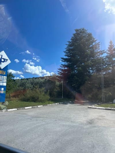 The image depicts a view from a vehicle, likely a car, looking towards a fuel station in Beloslav, Bulgaria. A tall, blue sign with a white logo and prices for fuel is visible on the left. The prices listed are "DIESEL 1.58" and "PETROL 1.54". Trees, including large evergreen trees, and vegetation are prominent in the background, with a hill covered in trees and shrubs behind them. The sky is bright blue with scattered white clouds, and the sun is shining, causing some lens flare in the upper right portion of the image. The foreground shows a paved area, likely part of the fuel station's forecourt, with a white and black kerb. The overall scene suggests a sunny day at a rural or suburban fuel station.