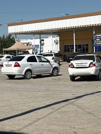 The image shows a street scene in the city of Pakhtakor, Uzbekistan. In the foreground, a white Chevrolet Aveo sedan is parked. The car has a license plate that reads "25R 121 FA". To the right of this car, another white sedan, possibly a Daewoo Nexia, is parked with its trunk open. Its license plate reads "25 U 190 JA". In the background, there is a building with a covered walkway supported by white pillars. A brown awning is set up under the walkway, and several people are visible under it, some appearing to be vendors or customers. A sign on the right side of the building displays text, some of which is legible as "AVIACIHI", "TEMIRYO'L", "DUBAY", and "AVTO SO'Z". The ground is paved with asphalt, and shadows suggest it is a sunny day. A tree is visible to the left of the building.