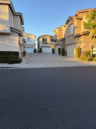 The image depicts a residential street in Aliso Viejo, California, USA, bathed in the warm glow of late afternoon sunlight. Several multi-story houses with beige and tan stucco exteriors line the street. Each house features a prominent garage with white overhead doors. The foreground of the image is dominated by a dark asphalt road. Beyond the road, a concrete sidewalk and driveway separate it from the houses. Landscaping consists of neatly trimmed green hedges planted along the base of the houses and in some areas beside the driveways. The sky is a clear, bright blue. A "NO PARKING FIRE LANE" sign is visible on the right side of the image, attached to a post near one of the houses. The overall scene suggests a quiet, suburban environment.