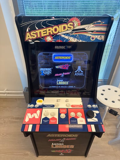 The scene depicts an Arcade1Up "Asteroids" arcade cabinet positioned indoors. The cabinet features a black body with multi-colored artwork. The top marquee displays "ASTEROIDS" in yellow lettering, set against a dark blue space background with stars, planets, a spaceship, and the Atari logo.

The machine's screen is illuminated, showing a game selection menu. Prominently displayed are the "Arcade1Up" and "ATARI" logos, along with game titles including "ASTEROIDS", "TEMPEST", "MAJOR HAVOC", and "LUNAR LANDER". Below the game titles, the instruction "PUSH A TO SELECT GAME ▼" is visible, and further down, "STRATEGY" text detailing gameplay mechanics and point values (20, 50, 100, 200 points) for different objects.

The control panel is designed with red, white, and blue segments. It features white buttons for "ONE PLAYER START", "TWO PLAYER START", "ROTATE LEFT", "ROTATE RIGHT", "THRUST", "FIRE", and "HYPER SPACE". A black trackball controller is located on the right side. Above these, there is an "OFF/ON" switch and "VOL+" and "VOL-" buttons. A white circular button on the left has a yellow sticker adhered to it, showing "1E33" and "£00" printed upside down. The lower cabinet side art repeats various game titles like "ASTEROIDS", "MAJOR HAVOC", "LUNAR LANDER", and "TEMPEST", along with the Atari logo.

The arcade machine is positioned in a room with a light-colored wooden floor. To the left, a plain light-colored wall is visible, with a grey radiator or heating unit partially seen near the floor. Behind the machine, a large window fitted with horizontal white Venetian blinds is present. Water droplets or condensation are visible on the exterior surface of the windowpane, suggesting precipitation. Natural light enters the room through this window. To the right of the arcade machine, a white stool with a circular, perforated seat stands on the floor. A black power adapter or electronic device is visible on the floor beneath the stool. No individuals are present in the image.

The scene is located in the city of Espoo, Finland.