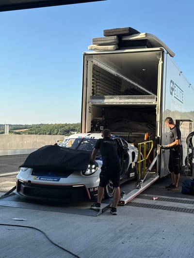A white racing car is being worked on, partially covered with a black cloth, inside a truck. Two men are visible, one near the front of the car and another deeper inside the truck. The truck's interior reveals metal racks and equipment. The truck has "Elite" written on its side. Outside, the sky is blue, and trees are visible in the background. The image is rotated 90 degrees counterclockwise.