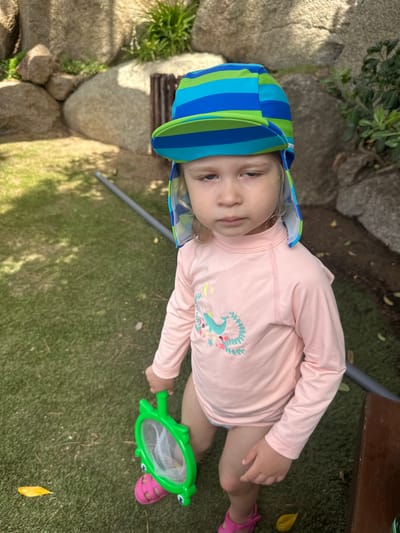 A young child wearing a pink long-sleeved rashguard with a whale design and a blue and green striped sun hat stands on a patch of artificial turf. The child holds a green turtle-shaped net in their right hand and is wearing pink sandals. In the background, large rocks and some greenery are visible, suggesting an outdoor setting, possibly a garden or a playground area. A gray pipe or rod runs across the ground behind the child. The lighting indicates it is daytime, with shadows cast on the grass. The child's expression is neutral. The scene appears to be located in Sant Feliu De Guíxols, Spain, given the context.