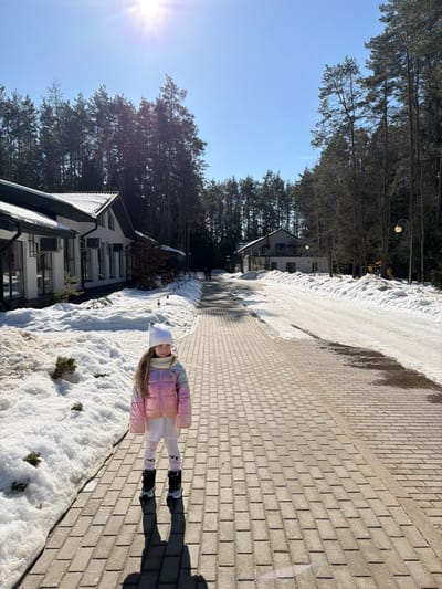 A daytime outdoor scene in Ракаўскі сельскі Савет, Belarus, features a young child standing on a paved pathway. The child, positioned in the foreground and facing the viewer's left, is dressed in a white knit beanie with cat ears, a light-colored knit scarf, an iridescent pink and yellow puffer jacket, white leggings adorned with a red pattern, and black and white winter boots. Her shadow is cast directly behind her on the light brown, brick-patterned pavers.
To the left of the pathway, several modern white buildings with dark sloped roofs are partially surrounded by snowdrifts and small evergreen shrubs. The closest building exhibits two air conditioning units mounted on its exterior wall. The paved path recedes into the distance, where two indistinct human figures are faintly visible in the midground. To the right of the pathway, a wider snow-covered area, mixed with exposed ground, extends towards another white building and a street lamp with a spherical light fixture. A dense forest of tall pine trees forms the backdrop on both sides and behind the structures. The sky is clear and bright blue, with the sun prominently visible in the upper-left quadrant, creating a lens flare and casting distinct shadows on the partially snow-covered ground.