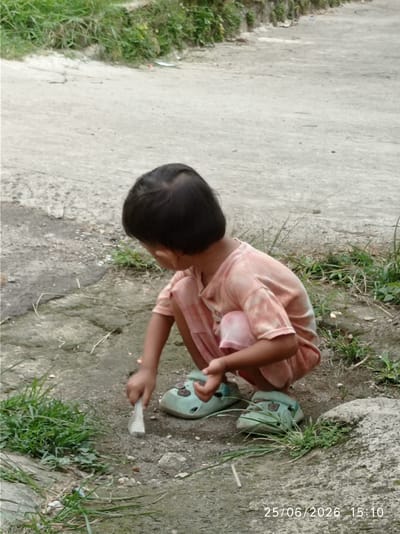 This media file shows a young child with dark hair, wearing a light peach-colored tie-dye t-shirt and matching shorts, crouching on the ground. The child is wearing light green clogs. The child is holding a light-colored object, possibly a stick or a tool, and appears to be digging or playing with the dirt. The setting is outdoors, with a paved surface that appears to be a path or road in the background, bordered by green grass and some rocks. The ground the child is on is a mix of dirt and small stones. The lighting suggests it is daytime. The image is dated 25/06/2026 15:10. The location context is Sukaraja, Indonesia. There are no strong emotional cues visible.