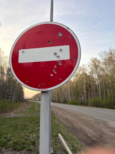 A road sign riddled with holes stands near a two-lane asphalt road bordered by trees. The sign is a red circle with a white horizontal bar across the middle, indicating "No Entry." It is mounted on a gray metal post. The sign shows numerous holes, suggesting it has been struck by bullets or projectiles. The edges of the road are lined with vegetation and patches of green grass. In the background, more trees and a glimpse of the horizon under a light sky are visible. The location is likely in Таскино, Russia.