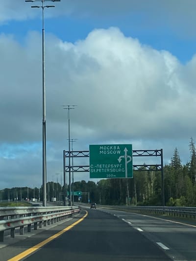 A green highway sign is positioned above a road in Tarasovo, Russia. The sign indicates directions to Moscow and St. Petersburg. The road ahead curves to the right, with a few cars visible in the distance. The environment is characterized by a cloudy sky, trees lining the side of the road, and a guardrail separating lanes. Tall streetlights are also present along the highway.
