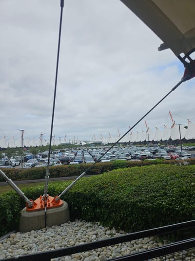 This image depicts a large, partially filled parking lot in Ashford, United Kingdom, likely near a retail or leisure complex given the presence of distinctive sail-like white canopies and building structures in the background. The foreground features a robust structural element supporting large cables, which are anchored to a concrete base covered with white pebbles. Behind this, a meticulously maintained hedge forms a boundary to the parking area. The parking lot itself is occupied by a variety of cars, and there are some people visible walking or standing among them. The sky is overcast with a mix of grey clouds and patches of lighter sky, suggesting a temperate climate. The overall scene conveys a sense of a modern commercial area on a cloudy day.