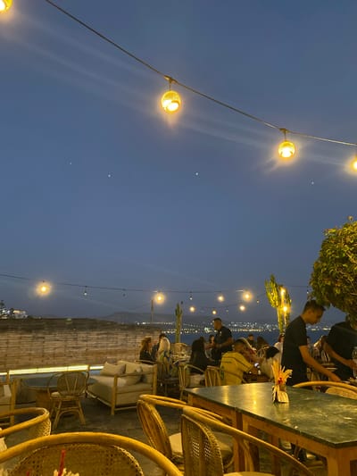 This is an outdoor evening scene at a restaurant overlooking the ocean in Taghazout, Morocco. The sky is a deep blue, transitioning to a lighter hue near the horizon, suggesting dusk. String lights with decorative spherical lanterns are suspended overhead, casting a warm glow. In the foreground, several wicker chairs surround a dark green, possibly marble-topped table. Beyond this, a low, cushioned seating area is visible, with more wicker chairs and tables. The middle ground shows a gathering of people seated at tables, some appearing to be dining or socializing. Two men in dark shirts are tending to tables, possibly serving. In the background, the lights of a city or town twinkle along the coastline, with the dark expanse of the ocean visible behind it. A low stone wall runs horizontally across the mid-ground, with a rustic wooden fence or partition above it. The overall ambiance is relaxed and inviting, typical of a beachfront dining establishment.