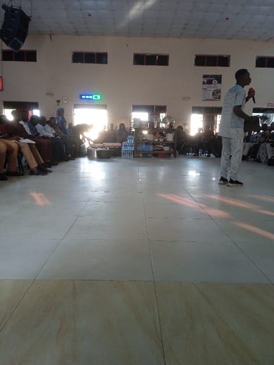 A medium shot captures a large room with rows of seated individuals on the left and right sides. A man stands on the right, holding a microphone and facing the group. Serving trays and water bottles sit in the center. The floor is tiled, reflecting sunlight streaming in through the windows. A clock hangs above a door, with speakers above the windows, and posters on the wall.
