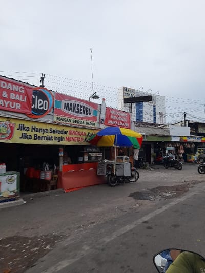 The image shows a street scene in Kecamatan Purwoasri, Indonesia, located at latitude: -7.6, longitude: 112.114. There is a food stall on the right side of the street, with a colorful umbrella and a red sign that reads "MAKSERBU" and "MAKAN SEBWA 10.000." The sign also has a slogan in Indonesian that roughly translates to "If you just want a snack, come to us. If you want to hang out, come to us." The street has a light gray asphalt surface and a white line down the center. The street is flanked by buildings with signs and awnings. On the left side of the image, a utility pole with many overhead wires is visible. There is also a blurry image of a motorcycle in the background, likely parked at the food stall. The sky is overcast and gray. The street appears to be relatively empty and quiet, with no people visible. The street is clean and well-maintained.