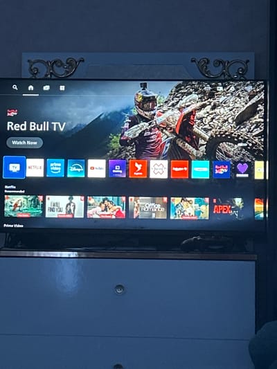 A television screen displays the Red Bull TV app interface. The screen shows a background image of a motocross rider in action, with a rocky, mountainous landscape behind them. The Red Bull TV logo and title are visible, along with a "Watch Now" button. Below this, a row of app icons includes Netflix, Prime Video, Disney+, YouTube, Sling TV, Xfinity TV, and Red Bull TV. Further down the screen, rows of recommended content are displayed, with titles such as "Netflix Recommended," "Prime Video," and "Netflix." The television is situated on a blue dresser with decorative metal trim on the wall above it. The overall scene suggests a home entertainment setup in Ognianovo, Bulgaria.