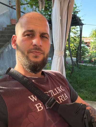 A portrait-oriented outdoor shot shows a man from the chest up, looking directly at the camera. He has a bald head, a full beard, and a mustache. He is wearing a dark red or maroon t-shirt with white text and a black sling bag across his chest, the strap clearly displaying "Reebok." A tattoo is visible on his right upper arm.
The background suggests a sunny outdoor setting, likely a garden or backyard in Rogozen, Bulgaria. Bright daylight illuminates the scene, casting highlights on the man's beard and face. Behind him, a beige or light-colored fabric canopy provides shade, supported by poles. Further back, lush green grass, trees, and bushes are visible. Some red flowers can be seen among the greenery. To the left, there are concrete or stone steps leading downwards, and a potted plant is partially visible. The overall impression is one of a relaxed moment in a casual outdoor environment.
Visible text includes "Reebok" on the bag strap and partial words on his t-shirt, such as "WRESTLING" and "RIA". The man's expression is neutral, looking steadily at the camera.