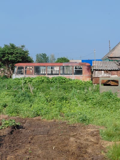 An abandoned, rusted bus sits in a grassy, overgrown area next to a dilapidated wooden structure. The bus, with its windows intact but frame visibly corroded, is partially obscured by tall green vegetation. To the right of the bus, a rustic wooden shed with a corrugated roof stands, appearing weathered and aged. The ground in the foreground is a mix of dark soil and dried grass. In the background, a clear blue sky stretches above a few scattered trees and some distant structures, suggesting a rural or semi-rural setting within the village of Novosyolovo, Russia. No people are visible in the scene.