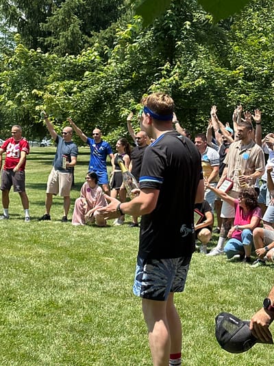 This is a medium-shot, vertically oriented photograph taken outdoors on a sunny day, likely in the late morning or early afternoon, given the strong shadows. The setting appears to be a grassy area with lush green trees in the background. A group of people, dressed in casual summer attire, are engaged in an outdoor activity or competition. They are standing in a line, with some individuals reaching out their arms.
In the foreground, a man wearing a black t-shirt with a blue and white striped collar and shorts with a palm-leaf pattern is prominent. He has a blue headband on. He is holding a bottle, possibly champagne, in his left hand. Behind him, several other individuals are visible, some holding what appear to be certificates or awards. One woman in a pink top and shorts is also reaching out. Further down the line, men in athletic shirts and shorts are participating. The man at the very bottom of the frame is wearing a red football jersey with the number 88.
The mood of the photograph seems to be energetic and celebratory. The people's postures suggest they are actively involved in a game or team-building exercise. The bright sunlight and green surroundings indicate a pleasant, warm weather event. The presence of awards suggests the conclusion of a competition or a significant moment in an event. The image's orientation suggests it might have been taken by someone standing or kneeling. The location context points to Razliv, Bulgaria.