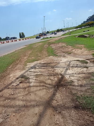 The scene depicts a roadside environment on a clear, sunny day in Karmajiji, Nigeria. In the immediate foreground, a broad, unpaved dirt path occupies the lower half of the image, characterized by uneven brown earth with interspersed patches of green grass. Distinct, dark shadows, consistent with overhead foliage or structures, are cast across the dirt. Moving towards the midground, a paved, multi-lane road is visible, bordered on its left by a continuous line of orange and white traffic barriers. Beyond the barriers to the left are additional green foliage and trees. On the right side of the road, a neatly maintained grassy embankment slopes upwards. Several vehicles are present on the road, including a dark sedan driving away from the viewer in the central portion of the main image and another dark vehicle further left. A single individual is observed walking on the far side of the road in the midground. The background is dominated by a bright blue sky with sparse white clouds. Numerous tall, slender streetlights line the road and grassy areas. In the central distance, several prominent, tall, white, sculptural structures stand vertically. Further to the right, a line of trees, some displaying red-orange blossoms, precedes a large, green-covered hill or small mountain in the far background.