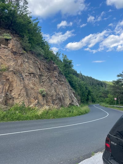 An image depicts a winding asphalt road passing by a steep, rocky cliff face covered in green vegetation. The road has a white dividing line. On the right side of the road, a black car with the word "GOLF" visible on its rear is parked partially in frame. A circular traffic sign with a red border and a white horizontal bar is also visible on the right. The background consists of a densely wooded hillside under a bright blue sky with scattered white clouds. The scene appears to be a natural landscape, possibly in or near Pechinska, Bulgaria, given the context provided.