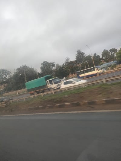 A slightly tilted photograph, exhibiting motion blur consistent with a capture from a moving vehicle, depicts a multi-lane highway in Biashara ward, Kenya, under an overcast sky. In the midground, a white truck with a green tarp-covered cargo bed is prominently visible, traveling from left to right. A white sedan, significantly motion-blurred, follows the truck. Further to the right, a yellow and blue bus is also present, blurred due to motion. The foreground consists of dark grey asphalt pavement, bordered by a brown earthen embankment with a black and grey concrete curb, topped by metal guardrails. The background features a dense line of various green trees and foliage, interspersed with a street lamp pole. Indistinct architectural structures are faintly discernible beyond the tree line. No human figures are explicitly visible.