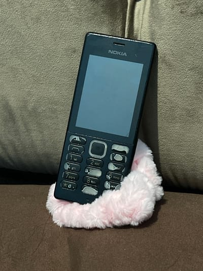 A black Nokia mobile phone is positioned on a light brown textured surface, possibly a sofa. The phone is resting on a soft, fluffy pink band, which appears to be a scrunchie. The phone's screen is dark, and the keypad is visible with numbers and letters. The background is a textured, muted green or gray fabric. The lighting suggests an indoor environment. The image is a close-up shot, focusing on the phone and the scrunchie. There is no indication of the city of Bandarbireuen, Indonesia, within the visual information provided.