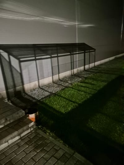 This is an image taken at night in the city of Pabradė, Lithuania. The scene depicts an outdoor area adjacent to a building with a grey, textured wall. A black, elongated canopy structure with a sloped roof casts a grid-like shadow on the wall and the ground. The ground below the canopy is covered in small, grey gravel, transitioning into a patch of dark green grass. A long, dark shadow from the canopy stretches across the grass. To the left, a set of outdoor stairs made of light grey interlocking bricks leads up to a paved area. Near the bottom of the stairs, a rusted, orange cylindrical container with a dark opening is visible on the grass. The lighting suggests artificial light sources, creating distinct shadows and highlighting the textures of the surfaces. There are no people or animals visible in the image.