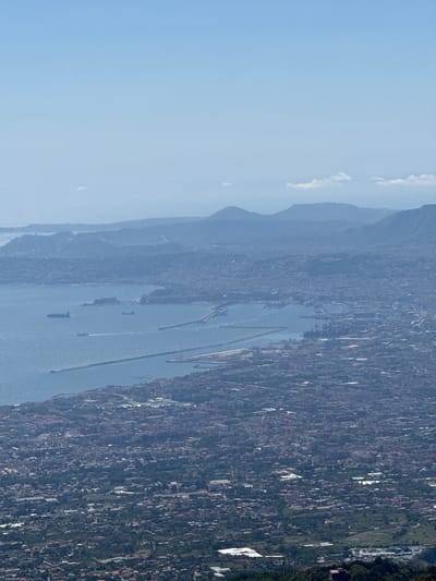 This is a high-angle aerial view of Ercolano, Italy, looking out over the Bay of Naples. The scene is dominated by the sprawling urban landscape of Ercolano, characterized by densely packed buildings with red-tiled roofs, interspersed with green patches of vegetation. The city extends to the coastline, where a large harbor and industrial area are visible. Numerous ships, including cargo vessels and smaller boats, are present in the water. The bay itself is a deep blue, reflecting the clear, pale blue sky. In the distance, a series of hazy, undulating hills and mountains form the horizon. The overall impression is of a vast, sun-drenched coastal city with significant maritime activity.