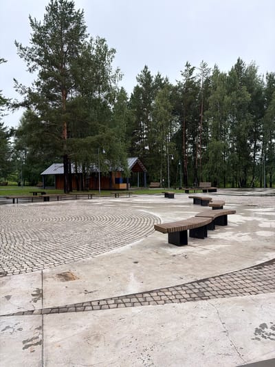 The image depicts an outdoor scene featuring a paved area with a curved design, bordered by cobblestones. Several modern, slatted wooden benches with black planters are strategically placed along the paved surface. In the background, a dense forest of tall evergreen trees creates a natural backdrop. Partially visible within the trees is a contemporary wooden building with a metallic roof, suggesting a park or recreational area. Streetlights are also present, hinting at an urban or semi-urban park setting. The ground appears damp, and the sky is overcast, indicating a cloudy or possibly rainy day. The image is taken from a low angle, looking upwards towards the trees. The orientation of the image is rotated 90 degrees counter-clockwise. The location is identified as Belebei, Russia. There is no visible text in the image.