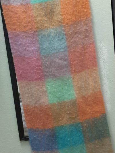 A multi-colored, patchwork textile, exhibiting a fuzzy or hairy texture, is depicted hanging vertically and slightly askew in an indoor setting within Shoreline, United States of America. The textile features a block pattern with squares and rectangles in shades of orange, pink, light blue, green, and brown. The textile partially obscures a black-framed object, possibly a mirror or picture frame, visible along the left edge of the image. The background consists of a light-colored wall, likely off-white or a light gray tone. No individuals are present.