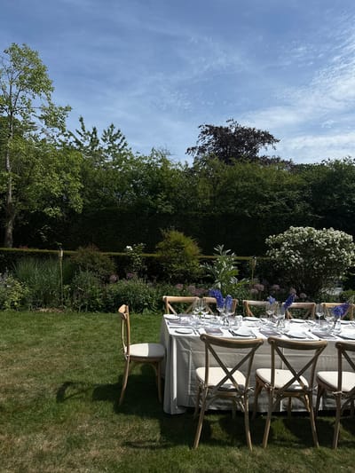 The image depicts an outdoor dining setup in a garden setting, likely in the city of Cambridge, United Kingdom. A long, grey-clothed table is arranged with place settings, including plates, glasses, and cutlery. Wooden chairs with off-white cushions are placed around the table. Small arrangements of blue flowers decorate the table's center. The dining area is situated on a lawn, bordered by various trees, hedges, and flowering plants. The sky is partly cloudy.