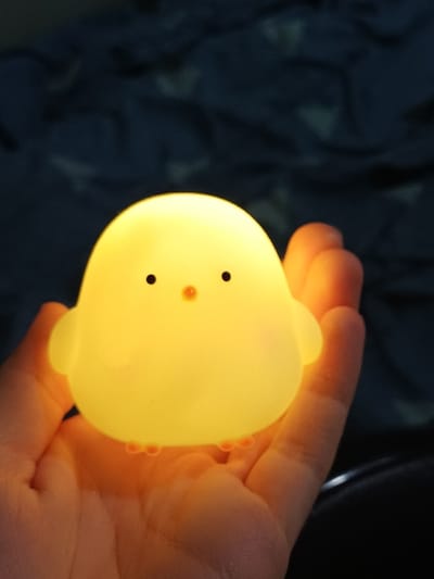 A luminous, yellow, chick-shaped night light is depicted resting within an open left hand. The object, composed of translucent material, emits a warm yellow glow and features two small black circular eyes and a minute orange dot for a beak. Small, bulbous orange feet are visible at its base. The hand, illuminated by the object's internal light, occupies the lower half of the frame. The background consists of dark, out-of-focus, rumpled fabric, consistent with a blanket or bedding. The scene is located within an interior space, likely a bedroom, in 서대문구, South Korea, under low ambient light conditions.