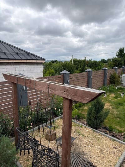 The image depicts a backyard scene in Semiluki, Russia. In the foreground, a wooden pergola supports two macrame swings hanging from metal chains. The swings are black and appear empty. To the right of the swings, a gravel area is visible, with some scattered stones and a wooden log. Beyond the pergola, a horizontal wooden fence with gray posts separates the yard from the surrounding area. The fence is adorned with string lights featuring clear bulbs. Behind the fence, lush green trees and bushes form a dense backdrop. A portion of a white brick house with a dark metal roof is visible in the upper left corner of the image. The sky is overcast with heavy, gray clouds. The overall scene suggests a residential outdoor space, possibly a garden or patio area.