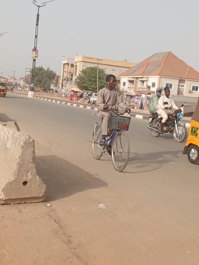 The image captures a sunny street scene in Sani Mai Nagge, Nigeria, depicting daily life and transportation.
In the foreground, a young man rides a bicycle. He wears a light-colored, long-sleeved traditional garment, possibly a caftan or jalabiya, and has white earphones visible in his ears. His bicycle has a front basket. Distinct shadows are cast on the light brown road surface by the cyclist and a concrete road barrier positioned on the left side of the frame, suggesting it is either morning or late afternoon.
To the right of the cyclist, two men are riding a motorcycle. The driver wears a light-colored outfit and a cap, while the passenger behind him is dressed in light green and wears a face mask. Partially visible on the far right is the front portion of a yellow commercial tricycle, often known as a 'keke napep', with the letters "KHC" visible on its side. In the far left background, another yellow tricycle can be seen.
The background features several buildings along the street. A prominent, light-colored building clearly displays the text "EXPRESS PLAZA." Next to it, another building has a distinct reddish-brown pyramidal roof equipped with several solar panels. Scattered trees provide some greenery along the roadside. A tall, modern-looking street lamp stands on the left. The sky is clear and bright, indicating good weather. The scene depicts people engaged in typical urban travel activities.