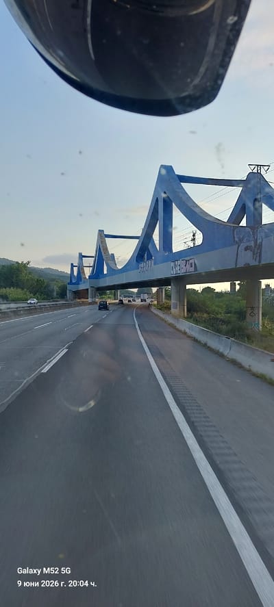The image shows a view from a vehicle driving on a highway in Llinars del Vallès, Spain. A distinctive blue bridge, with graffiti on its underside, spans the highway. The bridge has a modern, angular design with large triangular support structures. The highway itself has multiple lanes, with several cars and trucks visible in the distance. The sky is clear and blue, suggesting daytime. The lower part of the image contains a watermark indicating "Galaxy M52 5G" and the date "9 юни 2026 г. 20:04 ч.", which translates to June 9, 2026, at 8:04 PM. This watermark is likely from the device that captured the photo, not necessarily indicative of the actual time or date the photo was taken. The overall scene depicts a typical highway commute under a bright sky, with the blue bridge serving as a prominent landmark.