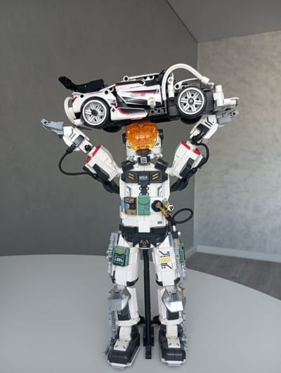 A white and black Lego robot stands on a white table, holding a white Lego car above its head with both hands. The robot is detailed, with a silver helmet topped with orange, and white and black armor plating. Small green and black compartments are visible on its torso, and a hose with a brass fitting connects to its chest. Text on the robot includes "DSA" on its chest, "201x" on a green compartment on its leg, and "EXO SKELETON" on its thigh. The car, also white and black, has a spoiler and racing stripes, with "RACING C" visible on its side. The background is a plain grey wall, suggesting an indoor setting. The lighting is even, indicating it's likely daytime, possibly in a room with artificial lighting or natural light from a window not in view. No specific weather cues are present. The scene is static, with the robot posed as if presenting or displaying the car. The overall impression is a clean, well-lit display of intricate Lego models.