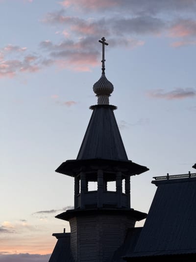 The image shows the top of a wooden church spire with a cross at its apex, set against a pale blue sky with wispy pink clouds. The spire is dark wood, with a tiered roof structure and a rounded, scaled dome just below the cross. To the right of the spire, a portion of another dark wooden roof with intricate decorative trim is visible. The lighting suggests either dawn or dusk. The scene is devoid of people. This architectural style is characteristic of churches in the Neman rural council, Belarus.
